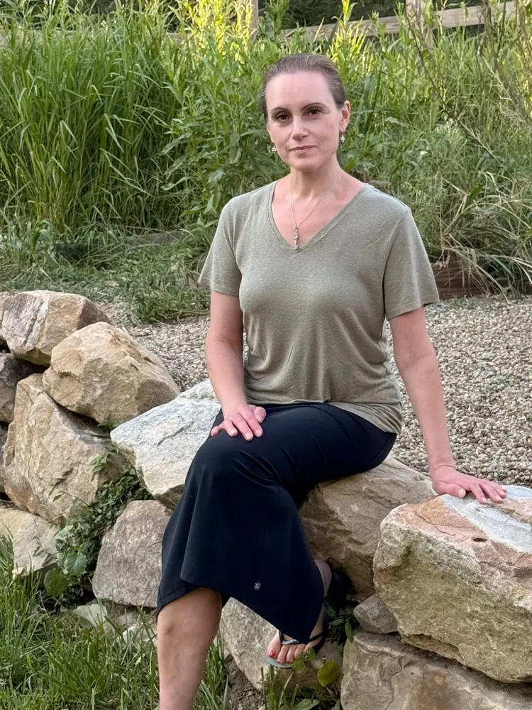 Anastassia Schlussel, owner of Alpha State Hypnosis, seated on a stone wall by the water